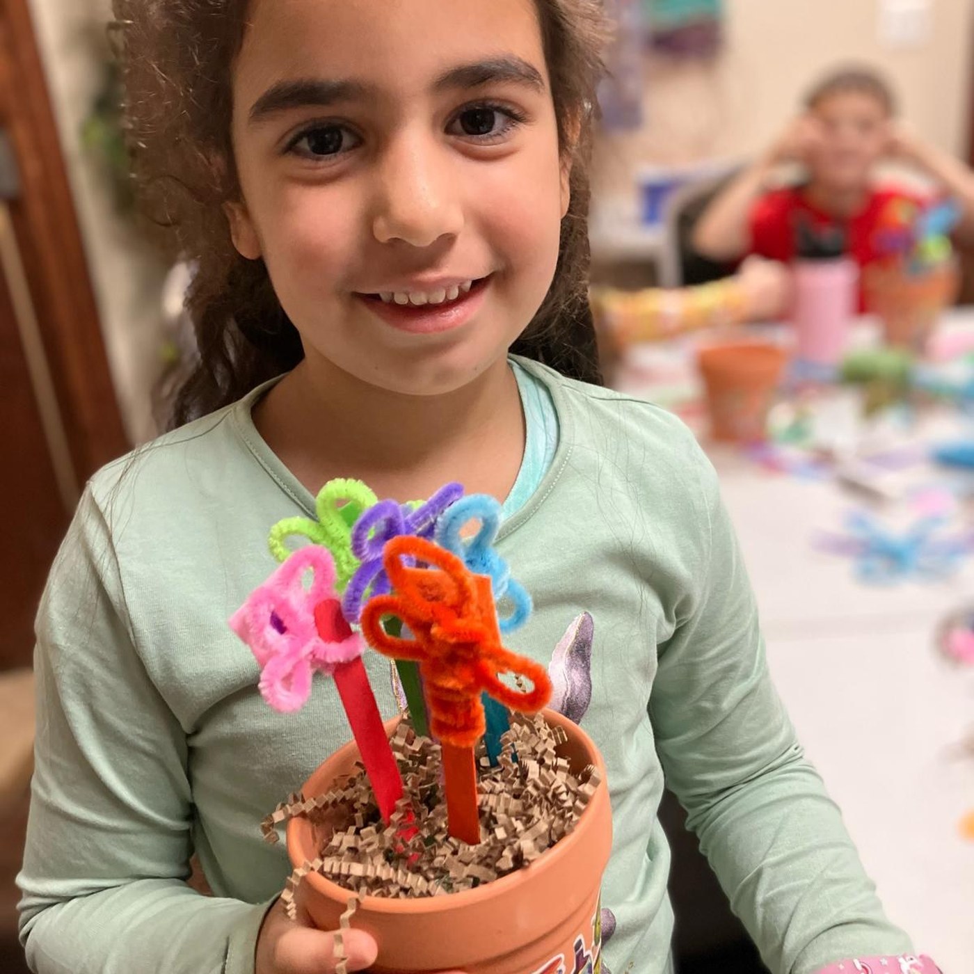 A child holding up a decorated flower pot she made, with classmates working at the table behind her.