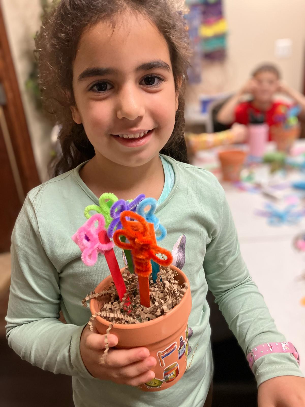 A child holding up a decorated flower pot, classmates working at the table behind.