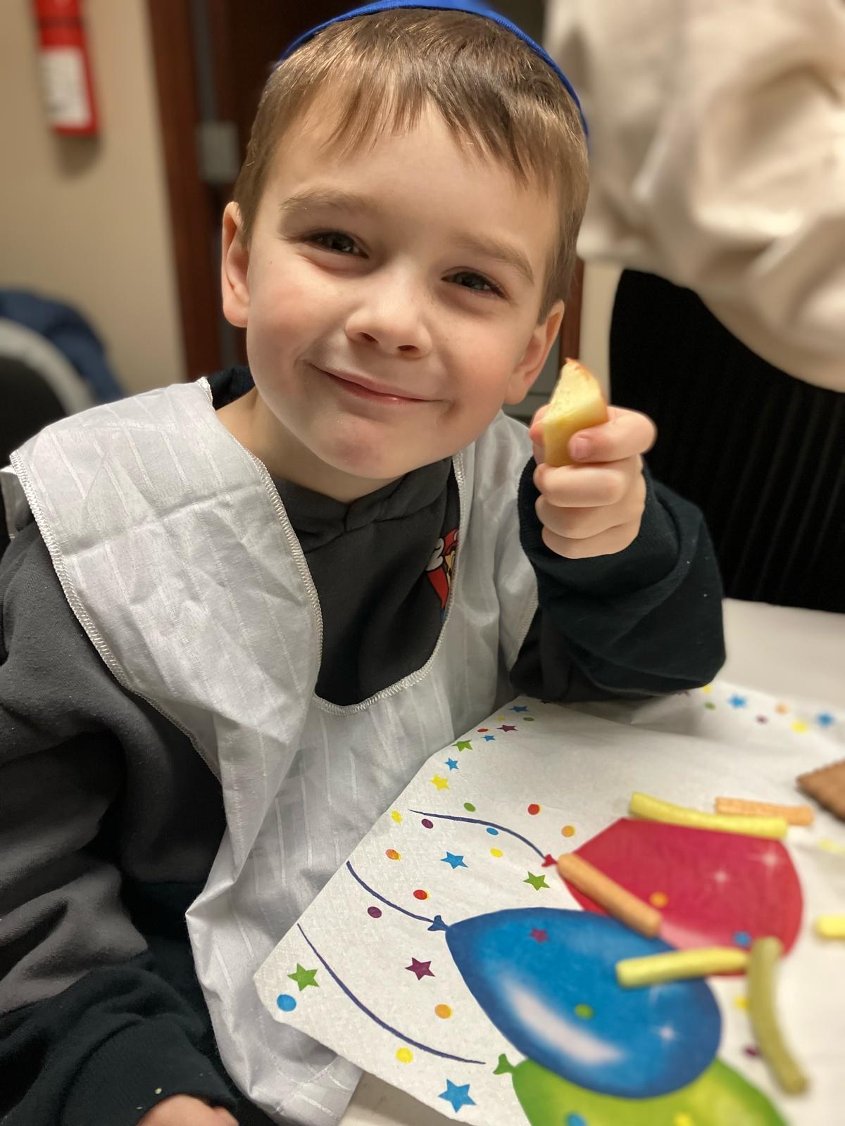 A child in a smock at the table, holding a piece of a snack.